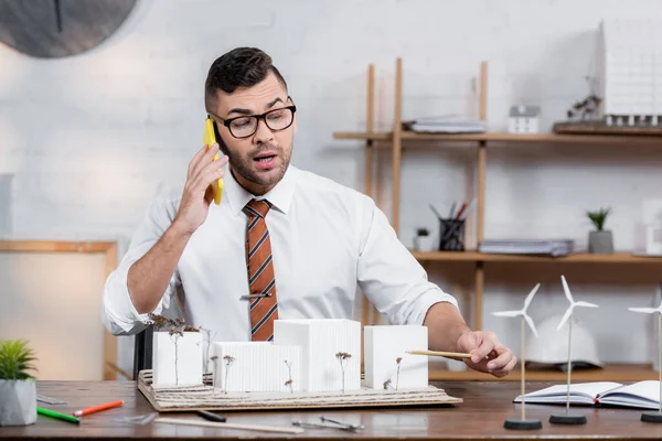 Architekt spricht am Arbeitsplatz mit Smartphone und zeigt mit Bleistift auf architektonisches Projekt — Stockfoto