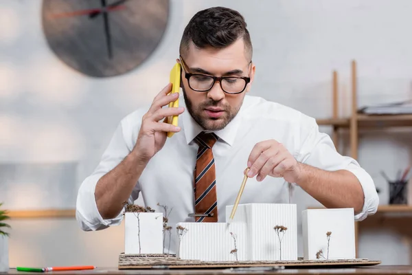 Architekt zeigt mit Bleistift auf Häusermodelle, während er am Arbeitsplatz mit dem Smartphone spricht — Stockfoto