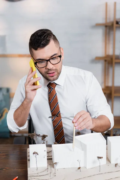 Architekt mit Brille spricht auf Handy und zeigt mit Bleistift auf Architekturprojekt — Stockfoto