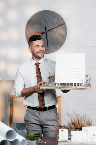 Architecte positif tenant maquette de bâtiment tout en se tenant debout sur le lieu de travail — Photo de stock