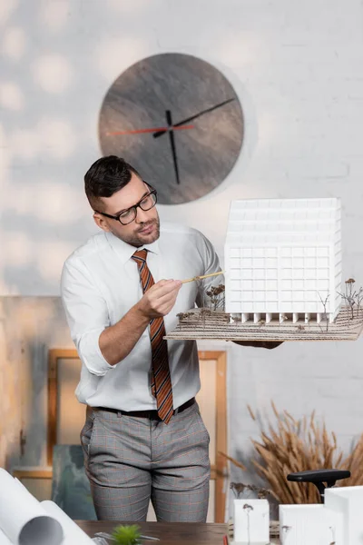 Architect in eyeglasses standing at workplace and pointing with pencil at house maquette — Stock Photo