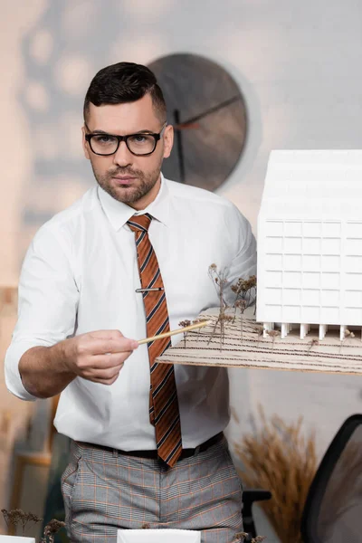 Serious architect looking at camera while pointing with pencil at house model — Stock Photo