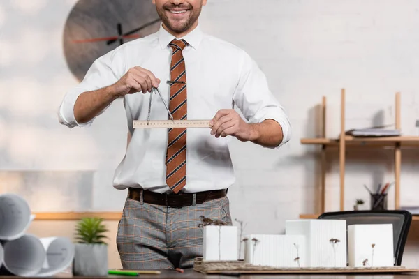 Vista recortada del arquitecto sonriente con regla y divisor cerca del proyecto arquitectónico, borrosa primer plano - foto de stock