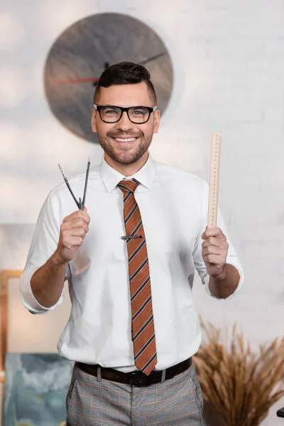 Cheerful architect in eyeglasses holding ruler and divider while smiling at camera — Stock Photo