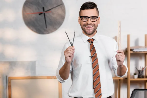 Happy architect looking at camera while holding ruler and divider in office — Stock Photo