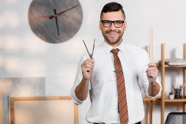 Arquiteto feliz olhando para a câmera enquanto segurando régua e divisor no escritório — Fotografia de Stock