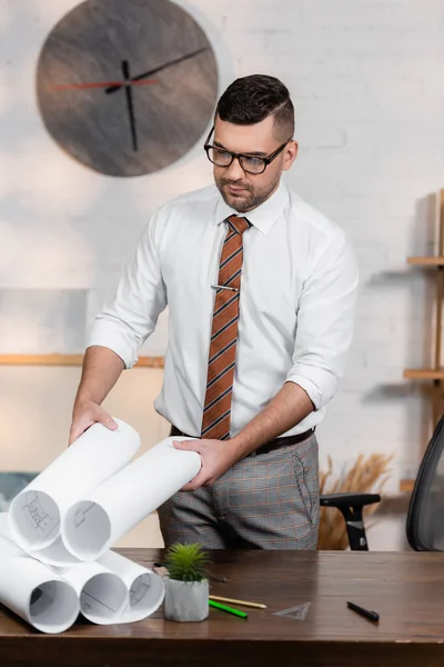 Architect in eyeglasses holding blueprint while standing at workplace — Stock Photo