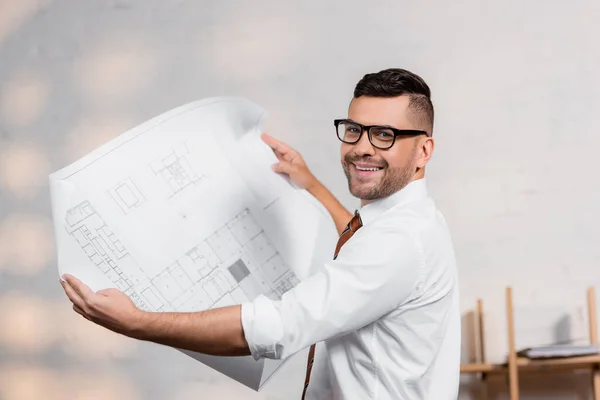 Arquitecto feliz en anteojos mirando a la cámara mientras sostiene el plano - foto de stock