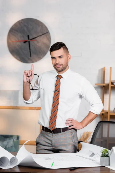 Arquitecto sonriente apuntando hacia arriba con el dedo mientras está de pie con la mano de la cadera cerca de planos — Stock Photo