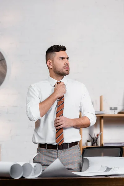 Seriöser Architekt schaut weg, während er Krawatte in der Nähe von Bauplänen im Büro justiert — Stockfoto