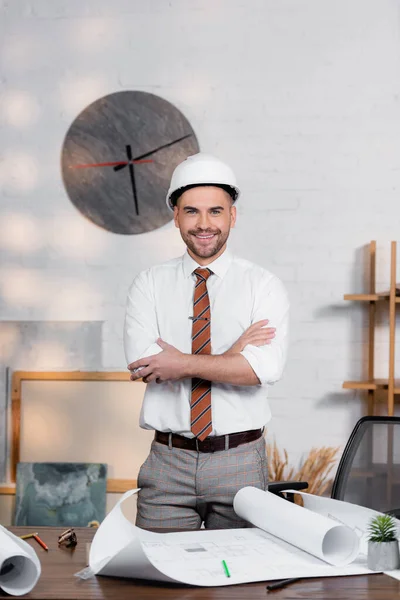 Щасливий архітектор у шоломі, що стоїть з схрещеними руками біля креслень на столі — стокове фото