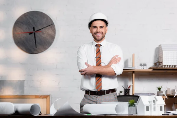 Lächelnder Architekt mit Helm, der mit verschränkten Armen in der Nähe von Bauplänen und Hausmodell steht — Stockfoto