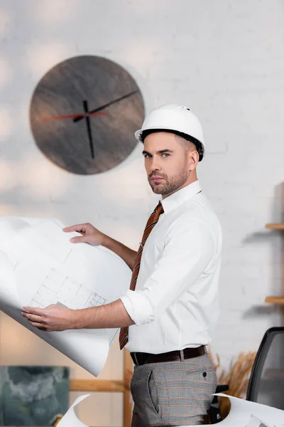 Serious architect in helmet holding blueprint and looking at camera — Stock Photo