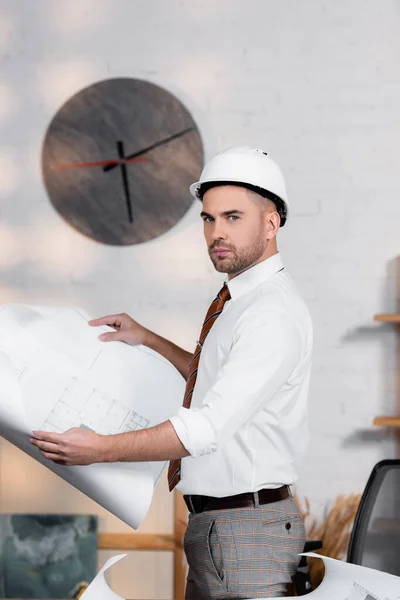 Arquitecto serio en casco sosteniendo plano y mirando a la cámara - foto de stock