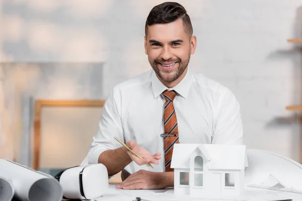 Arquiteto feliz olhando para a câmera enquanto aponta para o modelo da casa — Fotografia de Stock