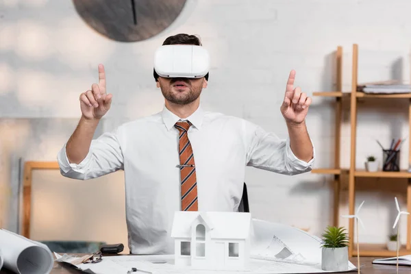 Arquitecto en auriculares vr apuntando con los dedos mientras está sentado en el lugar de trabajo cerca del modelo de casa - foto de stock