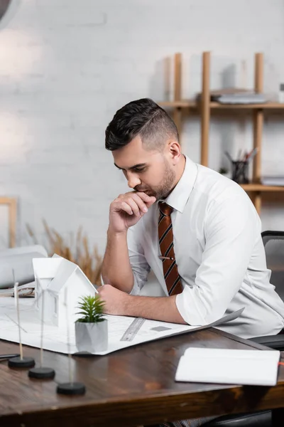 Thoughtful architect sitting at workplace near house model and blueprint — Stock Photo
