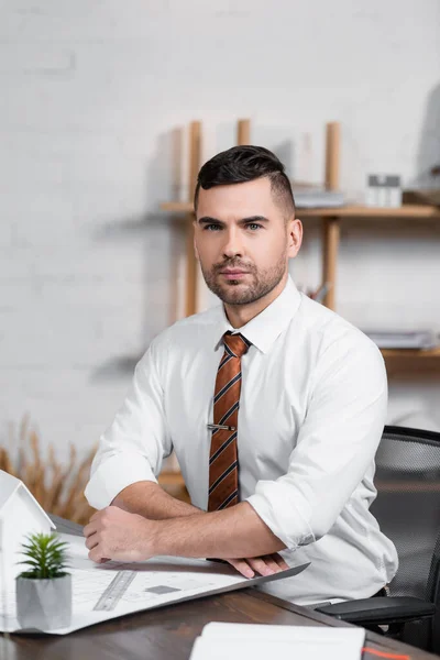 Serious architect looking at camera while sitting at workplace on blurred foreground — Stock Photo