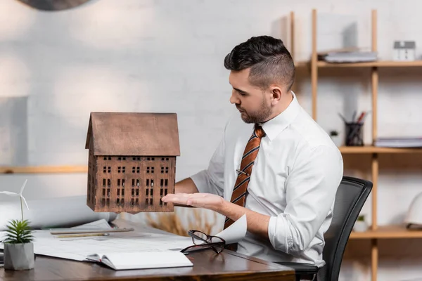 Architekt sitzt am Arbeitsplatz und zeigt mit der Hand auf Hausmodell — Stockfoto