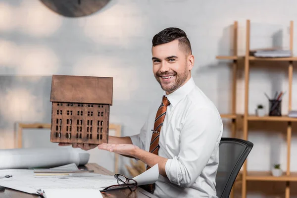 Glücklicher Architekt lächelt in die Kamera, während er auf das Hausmodell am Arbeitsplatz zeigt — Stockfoto