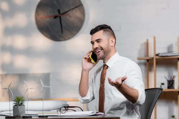 Arquitecto alegre hablando en el teléfono inteligente en el lugar de trabajo cerca del proyecto arquitectónico - foto de stock