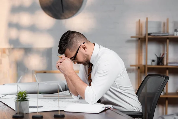 Müder Architekt sitzt mit gesenktem Kopf neben Bauplänen auf Schreibtisch — Stockfoto