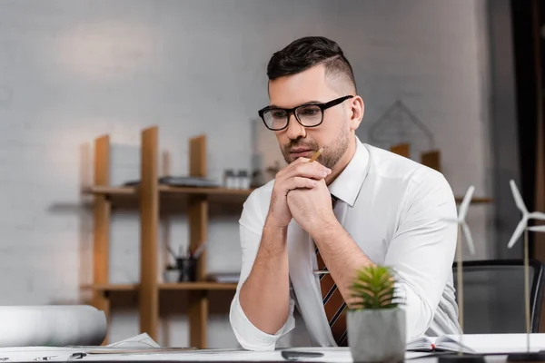 Thoughtful architect sitting at workplace near models of wind turbines on blurred foreground — Stock Photo
