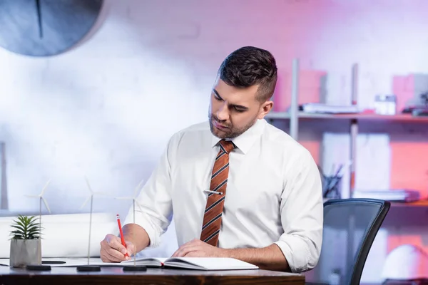 Concentrated architect writing in notebook near models of wind generators — Stock Photo