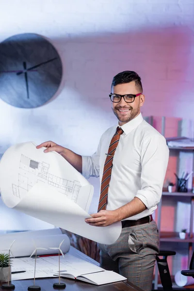 Arquitecto feliz mirando a la cámara mientras sostiene el plano cerca de los modelos de turbinas eólicas en el escritorio - foto de stock