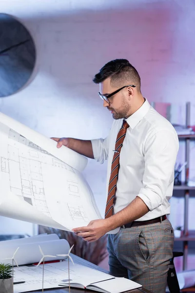 Serious architect looking at blueprint near models of wind turbines on desk — Stock Photo