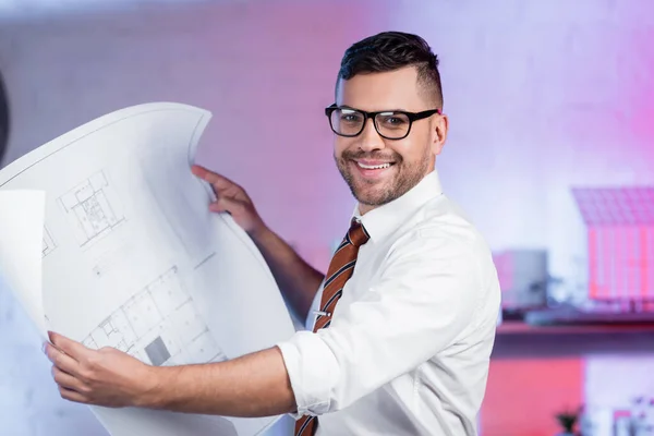 Happy architect in eyeglasses smiling at camera while holding blueprint — Stock Photo