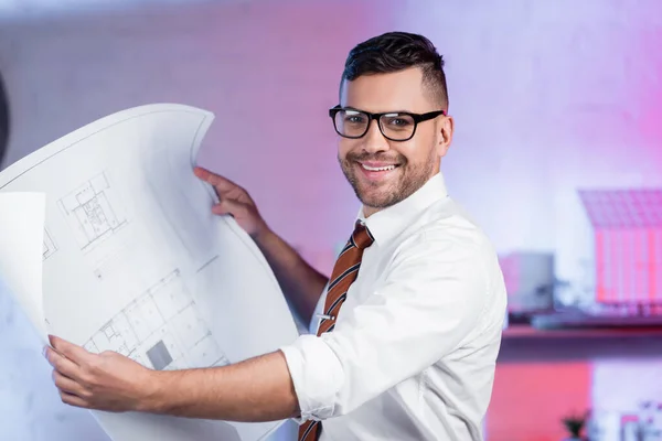 Feliz arquitecto en anteojos sonriendo a la cámara mientras sostiene el plano - foto de stock