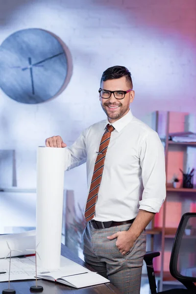 Happy architect standing with hand in pocket near rolled blueprint and smiling at camera — Stock Photo