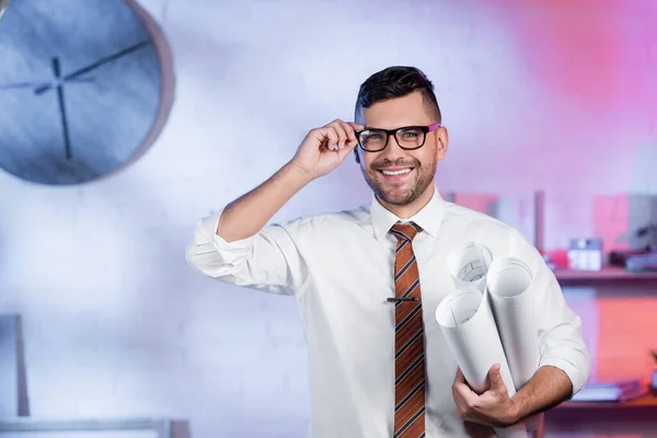 Arquitecto alegre sosteniendo planos enrollados y ajustando anteojos mientras mira la cámara - foto de stock