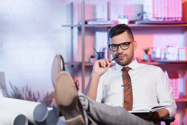 Positive architect looking at camera while holding notebook and pencil — Stock Photo