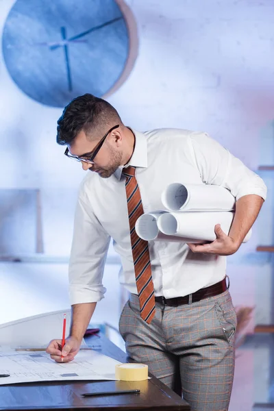 Architekt schreibt an Bauplan, während er Rollpläne im Büro hält — Stockfoto