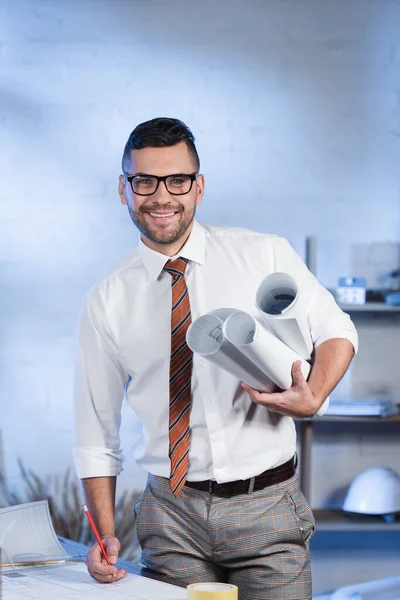 Cheerful architect with pen and rolled blueprints looking at camera in office — Stock Photo