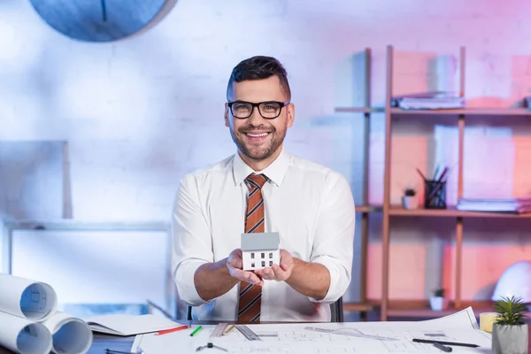 Zufriedener Architekt hält Hausmodell in der Nähe von Bauplänen auf Schreibtisch im Büro — Stockfoto