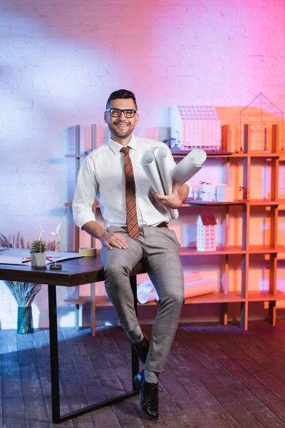 Arquitecto feliz sentado en el escritorio, sosteniendo planos enrollados y mirando a la cámara - foto de stock