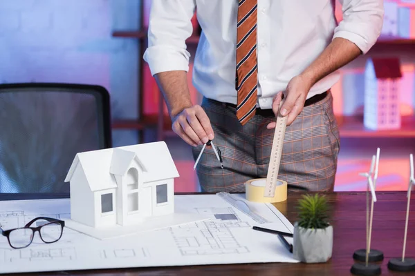 Vue partielle de l'architecte avec règle et diviseur près du modèle de la maison et le plan sur le lieu de travail — Photo de stock