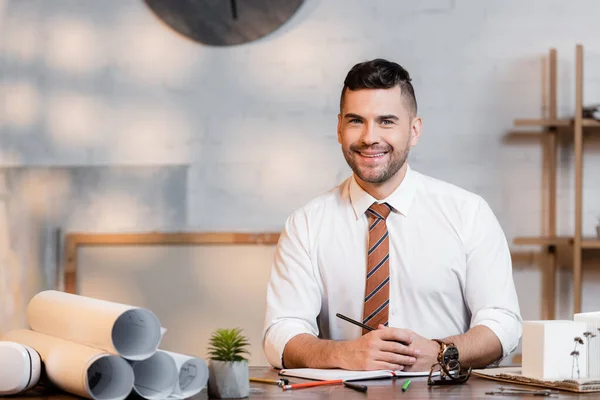 Smiling architect looking at camera while sitting at workplace near rolled blueprints — Stock Photo