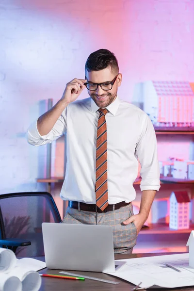 Lächelnder Architekt justiert Brille, während er mit der Hand in der Tasche am Arbeitsplatz steht — Stockfoto