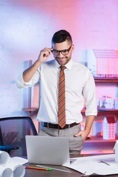 Architecte souriant ajuster les lunettes tout en se tenant la main dans la poche sur le lieu de travail — Photo de stock