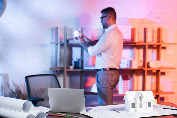 Enfoque selectivo de la computadora portátil, modelo de casa y planos en el escritorio, y arquitecto cerca de rack sobre fondo borroso - foto de stock