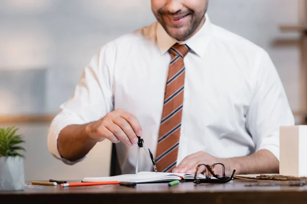 Vista recortada de arquitecto sonriente trabajando con divisor y cuaderno, fondo borroso - foto de stock