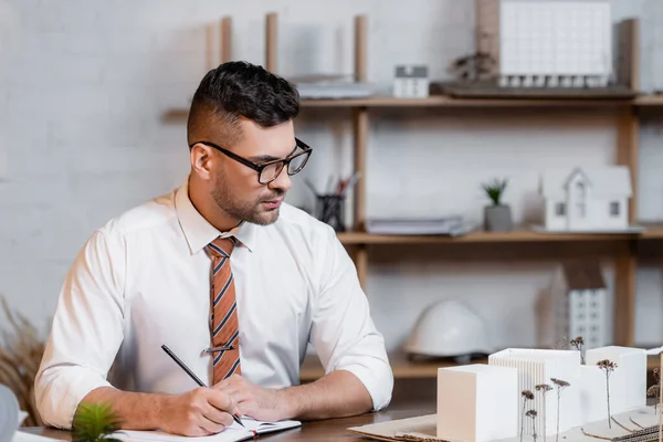 Architect writing in notebook near buildings models at workplace in office — Stock Photo