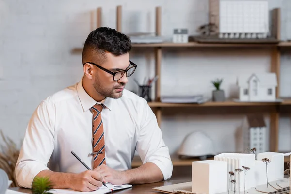 Architecte écriture dans carnet près de bâtiments modèles sur le lieu de travail dans le bureau — Photo de stock