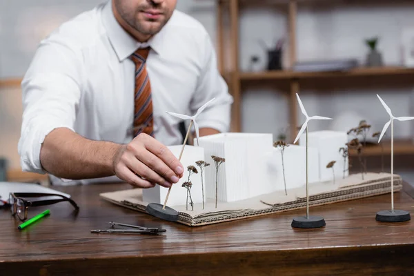 Cropped view of architect holding model of wind turbine near architectural maquette, blurred background — Stock Photo