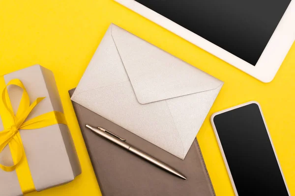 Top view of smartphone and digital device with blank screen on copy book with golden pen and envelope near gift box isolated on yellow — Stock Photo