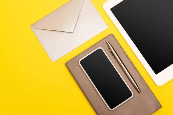 Vue du haut des appareils avec écran vierge près du bloc-notes avec stylo doré près de l'enveloppe isolée sur jaune — Photo de stock